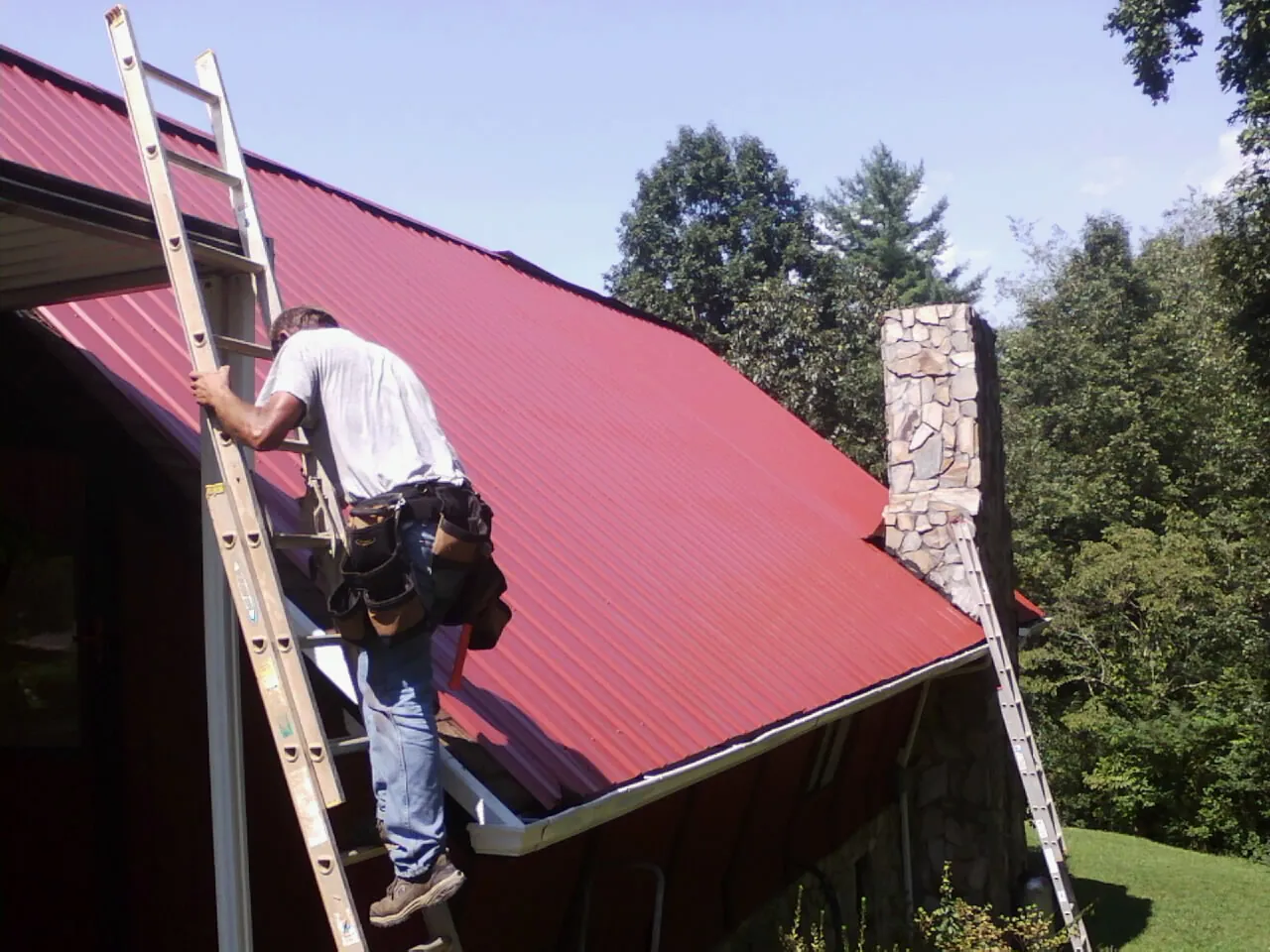 Roofer on a ladder beside a red metal roof and stone chimney — Soffit & Fascia Repair by Roofing Brothers in Glen Oaks